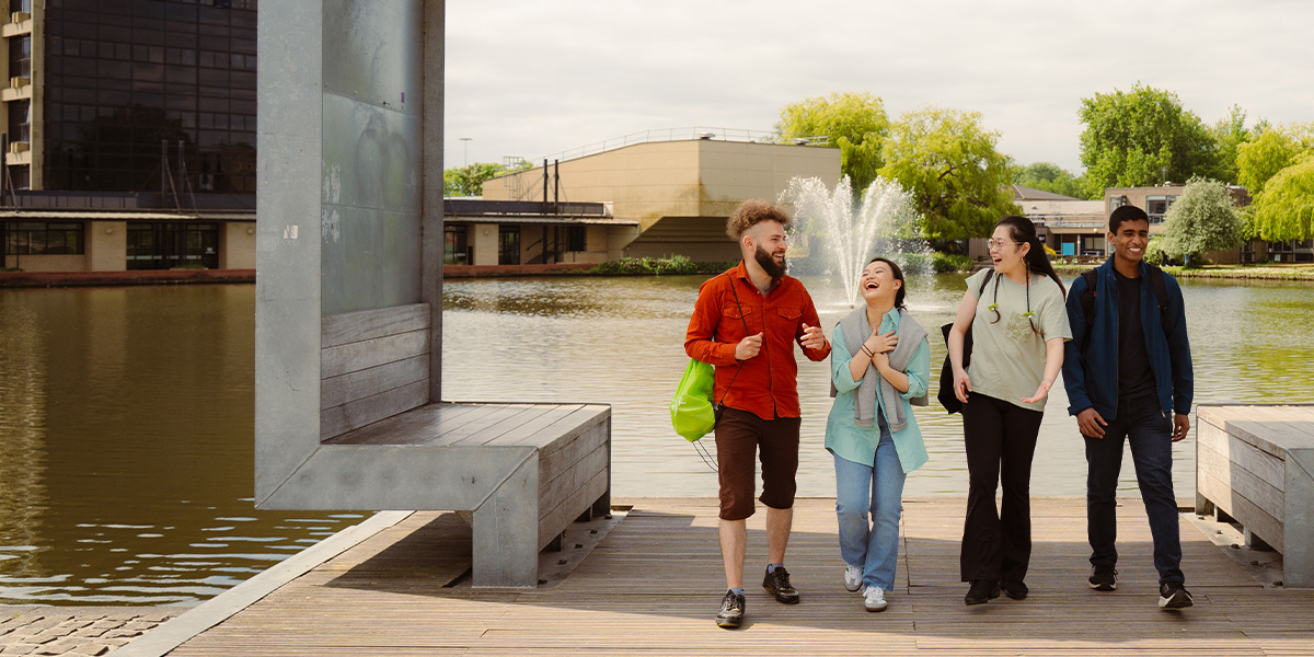 Four student chatting and laughing whilst walking away from the lake on Campus West.